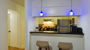 A compact kitchen with a central island, two bar stools, pendant lighting, and a view into the adjacent room.
