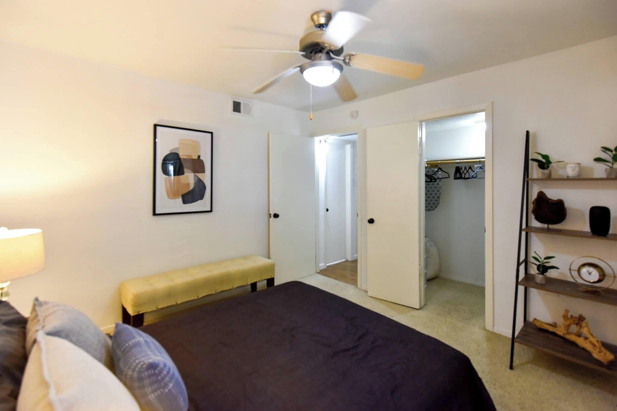A bedroom with a bed, a ceiling fan, a bench, a bedside table, a mirror, a closet, and a shelf with decorative items.