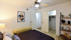 A bedroom with a bed, a ceiling fan, a bench, a bedside table, a mirror, a closet, and a shelf with decorative items.