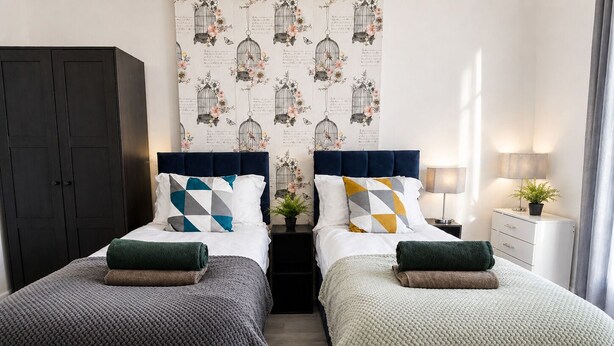 A double room with two beds, dark wooden wardrobe, patterned wallpaper, bedside tables, and a nightstand with a lamp. - Spacious Barnsley Home I Sleeps 6 I Parking I WiFi (Barnsley)