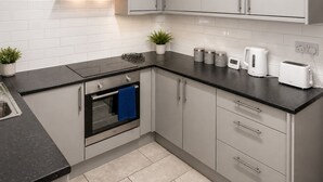 A modern kitchen with grey cabinets, a black countertop, a built-in oven, a kettle, a toaster, and a small potted plant. - Spacious Barnsley Home I Sleeps 6 I Parking I WiFi (Barnsley)