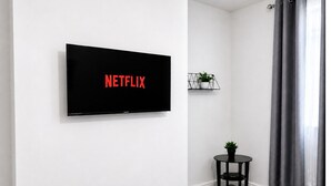 A room with a mounted TV displaying 'NETFLIX,' a small table with a plant, and grey curtains. - Spacious Barnsley Home I Sleeps 6 I Parking I WiFi (Barnsley)