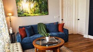 Un salon avec un canapé bleu, une table basse ronde, un pouf capitonné, un tapis à motifs et un tableau sur le thème de la forêt accroché au mur.