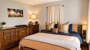 Une chambre avec un plancher en bois, un lit recouvert d’une couvre-lit beige et bleue, une commode en bois, un tableau accroché au mur et une fenêtre avec des rideaux.