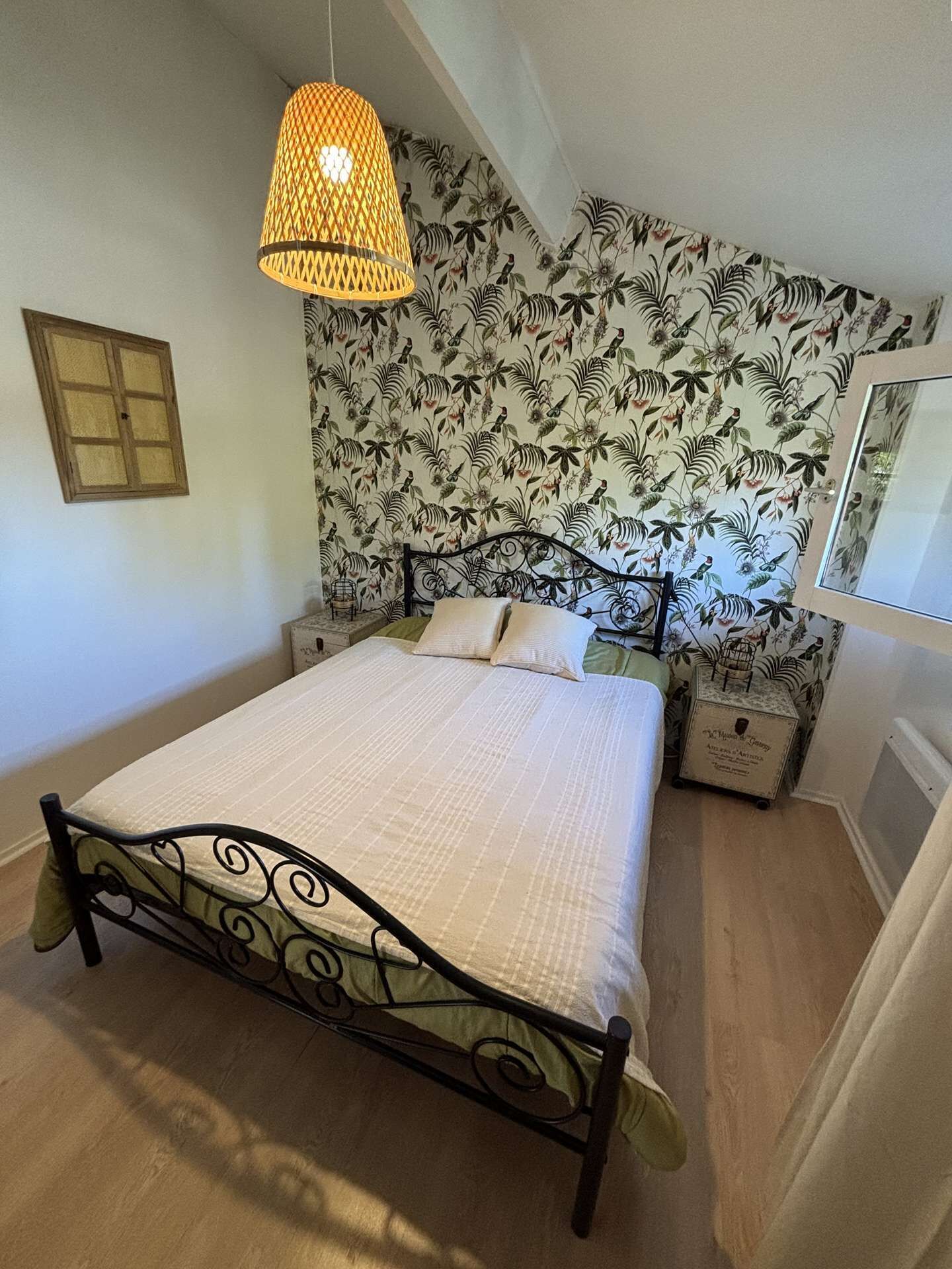 A bedroom with a bed, a hanging lamp, a floral wallpaper, a small bedside table, and a mirror.