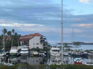 Ein Yachthafen mit vertäuten Booten, ein Gebäude mit Balkonen und ein Gewässer im Hintergrund.