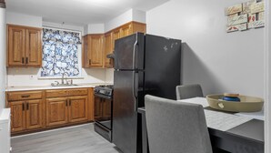 A kitchen with wooden cabinets, a black refrigerator, a dining table with a chair, and a window with a floral curtain.