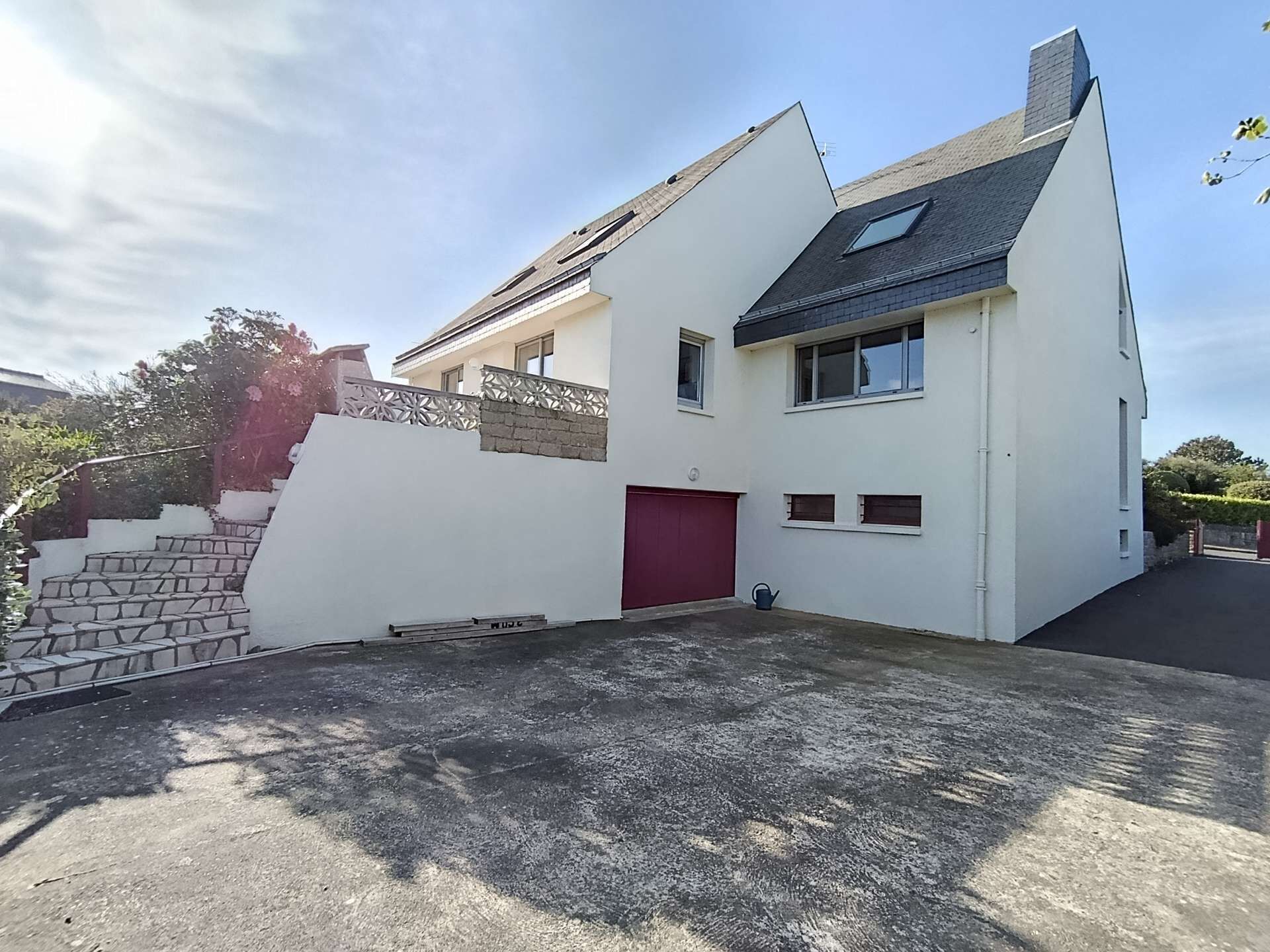 Une maison moderne à deux étages, dotée d’une porte de garage rouge, d’un toit en pente et d’une petite fenêtre à l’étage supérieur.