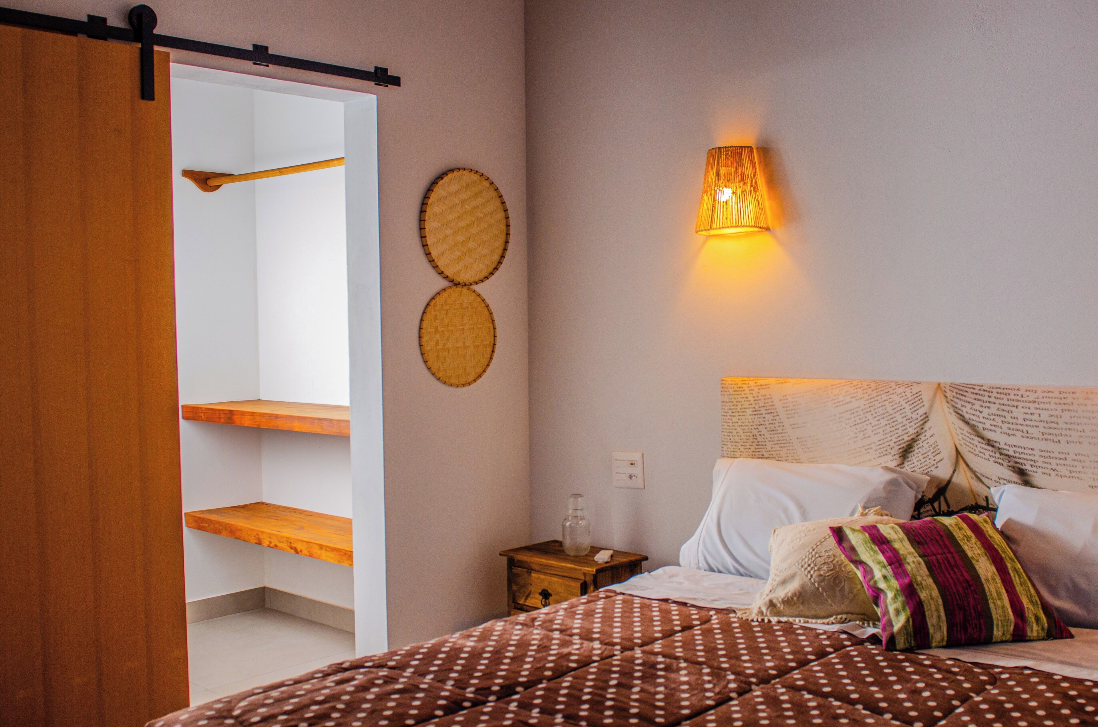 Un dormitorio con una cama, almohadas, una mesita de noche, una lámpara de pared y un estante de madera.