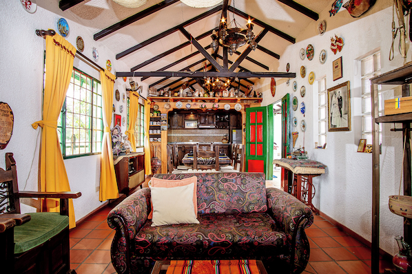 A cozy room with a patterned sofa, wooden beams, a chandelier, and a kitchen area.