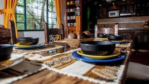 A traditional dining table with bowls and plates, a window with curtains, and a kitchen area with shelves.