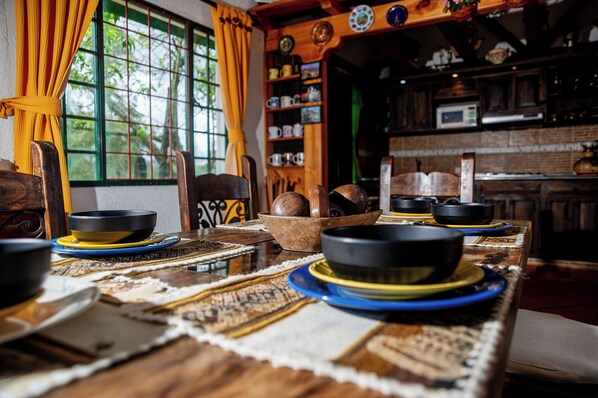 A traditional dining table with bowls and plates, a window with curtains, and a kitchen area with shelves. - Colonial-style house with a fireplace, 10 minutes from downtown. (Chiquinquirá)