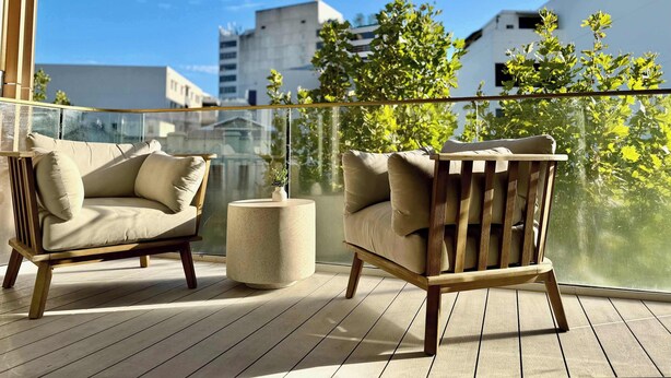 Een balkon met twee houten stoelen, een ronde tafel en uitzicht op gebouwen en bomen.