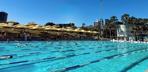 A swimming pool with multiple lanes, sun umbrellas, and a cityscape in the background. - Executive Designer Stay • Central • Gym • Sauna (Surry Hills)