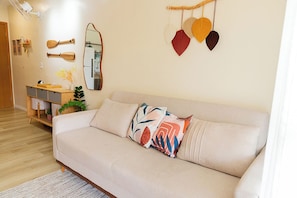 A living room with a beige sofa, a wooden side table, a mirror, and decorative items on the wall. - Beachfront apartment in Ilhéus - Vog Marine - Oceanfront pools - Ideal for families (Ilhéus)