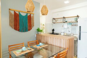 A modern kitchen with a central island, wooden chairs, a glass dining table, and hanging macramé wall art. - Beachfront apartment in Ilhéus - Vog Marine - Oceanfront pools - Ideal for families (Ilhéus)