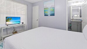 Un dormitorio con cama, televisor sobre una consola blanca, una puerta, un cuadro y un baño visible a través de una puerta entreabierta.