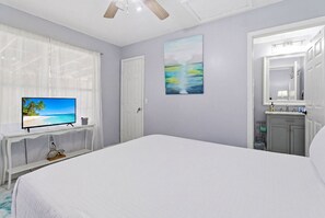 Ein Schlafzimmer mit einem Bett, einem Fernseher auf einer weißen Ablage, einer Tür, einem Gemälde und einem sichtbaren Badezimmer hinter einer geöffneten Tür.