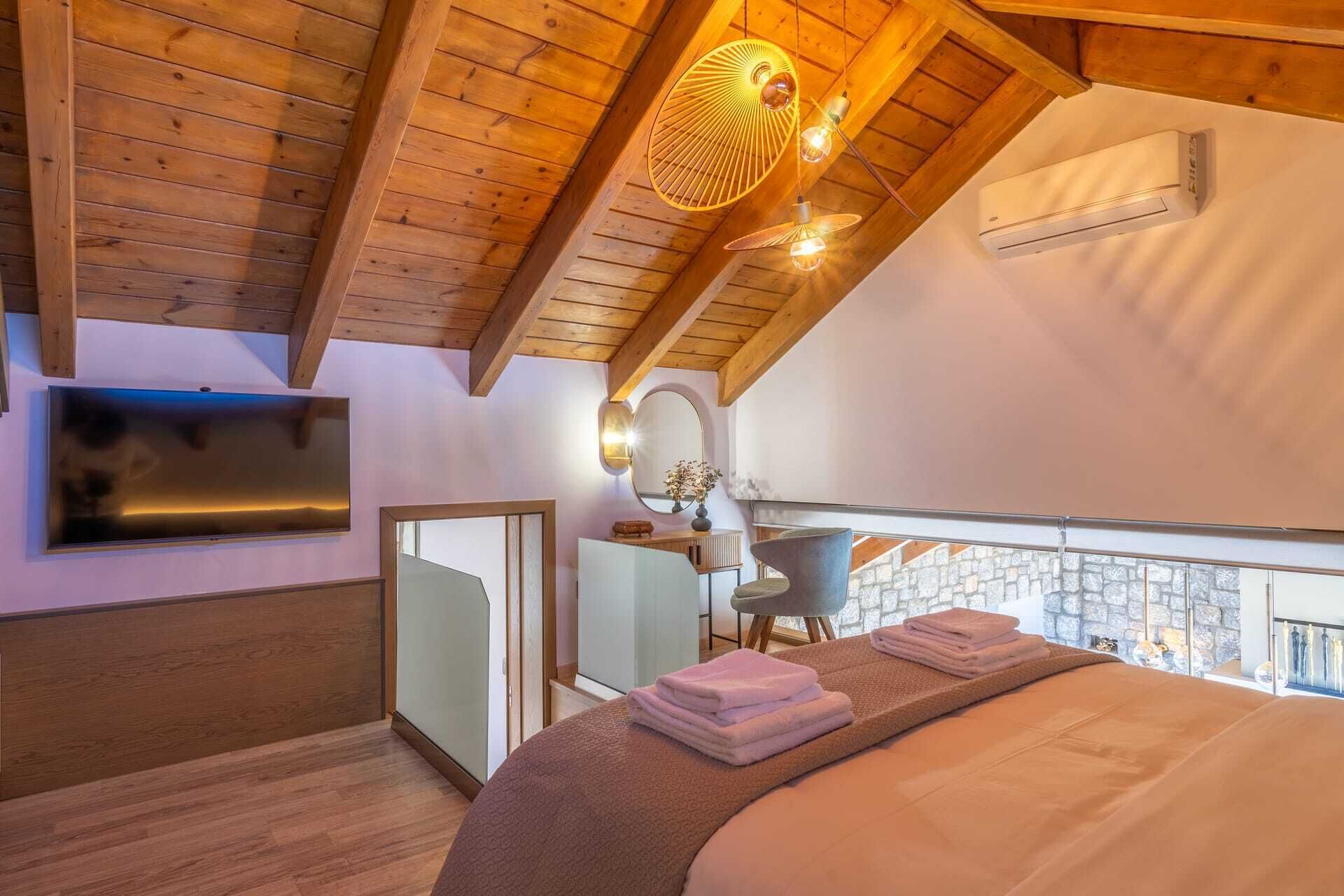 A bedroom with a sloped ceiling, a bed, a TV, a mirror, a chair, and a balcony with stone walls.