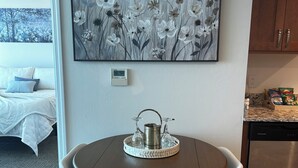 Uma sala de jantar com uma mesa redonda, duas cadeiras, um quadro na parede acima e uma cozinha ao fundo.
