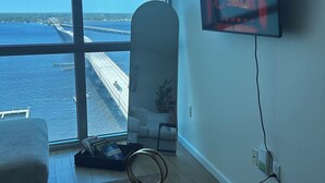 Quarto com uma grande janela com vista para a costa, uma televisão de tela plana na parede, uma mesa de centro de madeira e uma poltrona branca.