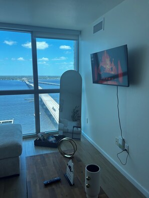 A room with a large window overlooking a coastal view, a flat-screen TV mounted on the wall, a wooden coffee table, and a white armchair. - Casa Privé-Amazing water view to relax and getaway (Ft Myers)