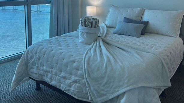 Quarto de hotel com uma janela ampla, cama com roupa de cama branca, abajur e um quadro na parede.