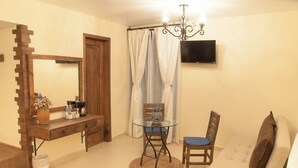 Una habitación con un escritorio de madera, una mesa de vidrio, una silla, un sofá, un televisor montado en la pared y una cortina.