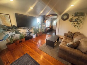 Ein Wohnzimmer mit Sofa, Couchtisch, Topfpflanzen, Fernseher und einer Küche im Hintergrund.