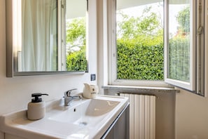 A bathroom with a white sink, a mirror, a window with a view of greenery, and a radiator.