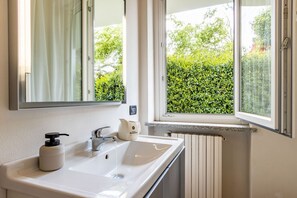 Un baño con un lavamanos blanco, un espejo, una ventana con vistas a la vegetación y un radiador.
