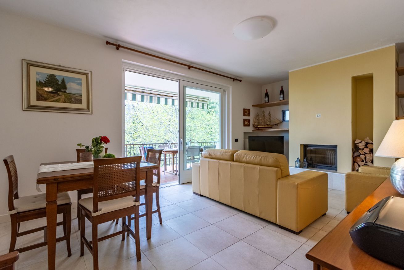 Una sala de estar con sofá, una mesa de comedor con sillas, chimenea y balcón con vista a árboles.