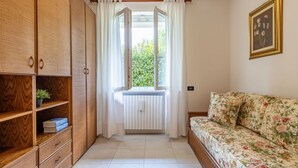 A room with a wooden wardrobe, a window with white curtains, a radiator, a floral sofa, and a framed picture on the wall.