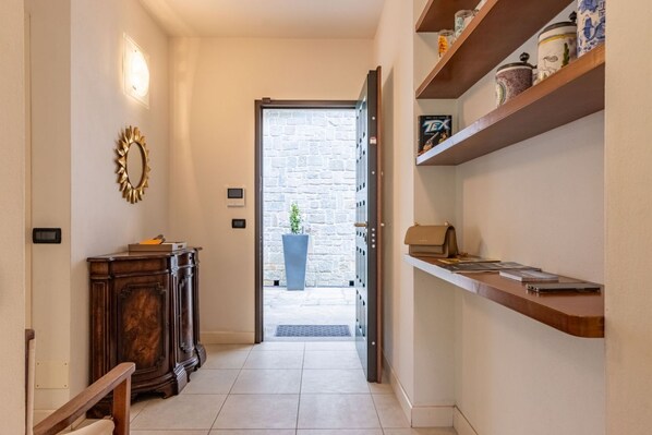 A hallway with a wooden cabinet, a mirror, a shelf with items, and a door leading to a stone wall.