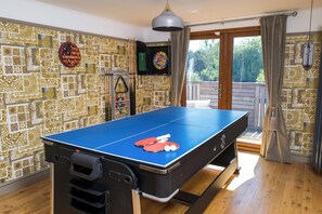 Une table de ping-pong avec raquettes et balles, un jeu de fléchettes et une bibliothèque dans une pièce dont les murs sont recouverts d’un papier peint à motifs et qui offre une vue sur l’extérieur.