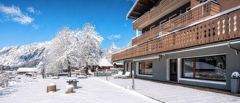 A modern building with a wooden balcony and large windows, surrounded by snow-covered trees and mountains.