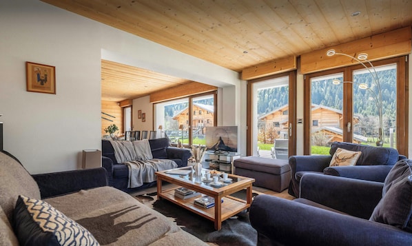 Un salon chaleureux avec un plafond en bois, de grandes fenêtres et une vue sur un chalet de montagne.