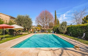 Une piscine rectangulaire avec une terrasse carrelée, entourée de verdure et avec un bâtiment visible à l’arrière-plan.