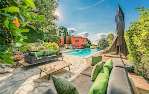 Ein Poolbereich mit einer Steinterrasse, Gartenmöbeln und Blick auf ein rotes Gebäude.