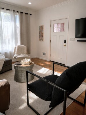 Ein Wohnzimmer mit einer Couch, einem Sessel, einem Hocker und einem Fernseher an der Wand.