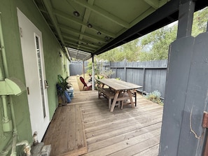 Une terrasse couverte avec une table et des chaises en bois, une porte avec une fenêtre, et une clôture en bois en arrière-plan.