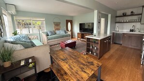 A modern living room with a sofa, TV, wooden table, and kitchen area with cabinets and appliances. - Hot Tub. A/C. Laundry. 2 Parking Spots. Orchard. (San Diego)