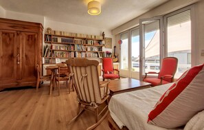 A cozy room with a wooden floor, a bookshelf filled with books, a red chair, a wooden table, and a bed with a white and red blanket.
