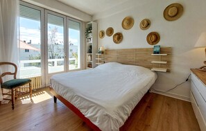 A bedroom with a large bed, a chair, a bookshelf, and a view through the window.