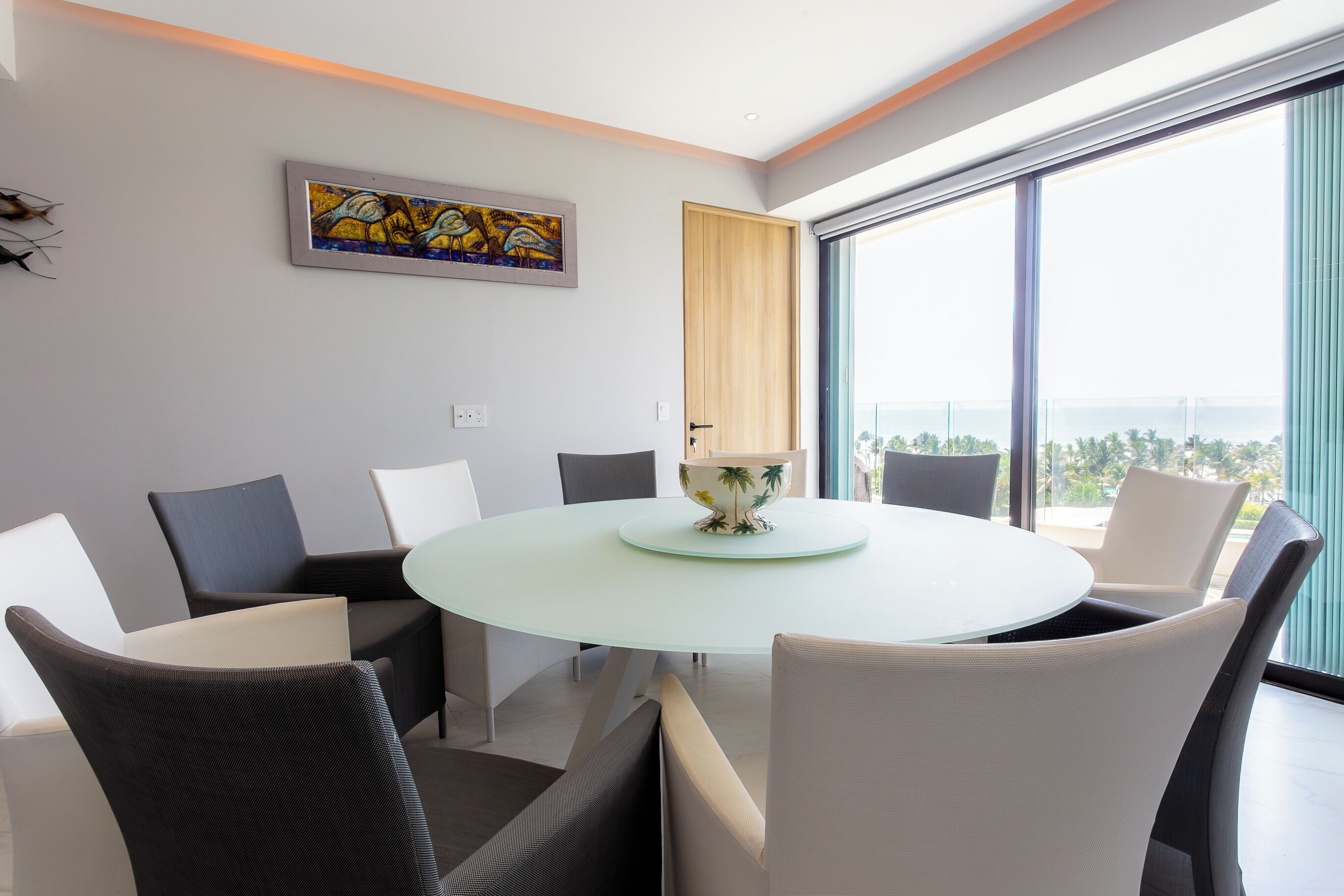 Een moderne eetruimte met een ronde glazen tafel, zes stoelen, een decoratieve schaal, een schilderij en een groot raam met uitzicht.