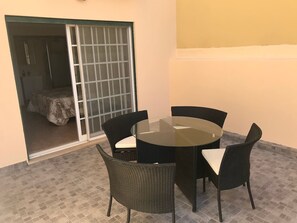 Ein Zimmer mit Glastisch und vier Stühlen, eine Schiebetür aus Glas, die zu einem Bett führt, und ein Fliesenboden.