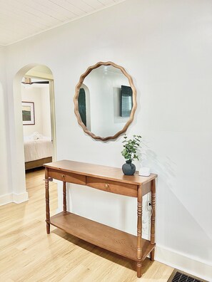 Une console en bois avec un miroir, un vase avec une plante et un lit en arrière-plan.