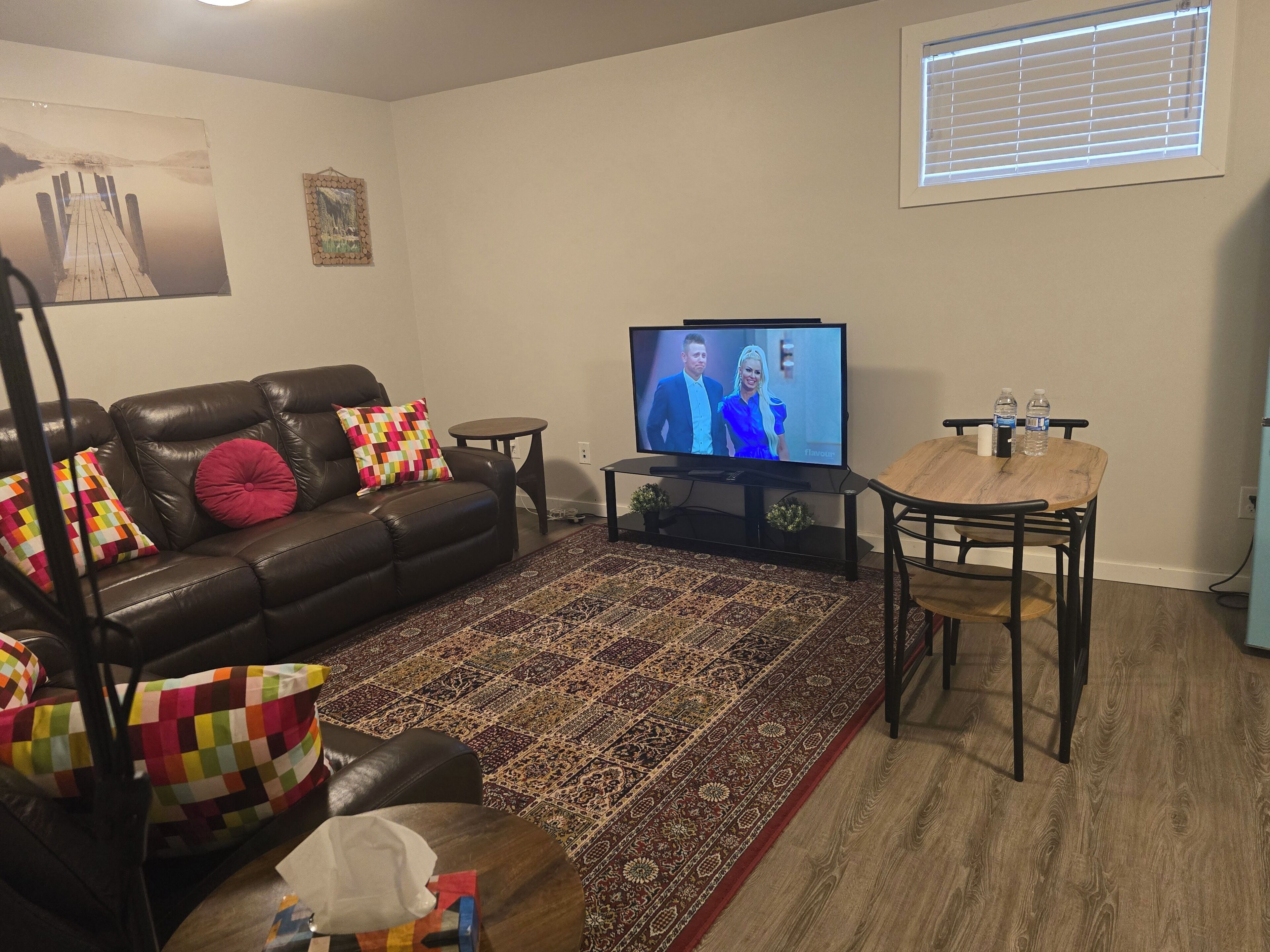 A living room with a leather sofa, a television, a patterned rug, a dining table, and a window with blinds.