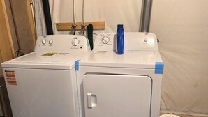 Een wasmachine en droger met bedieningsknoppen, een blauwe fles wasmiddel en een witte plastic zak op de vloer.