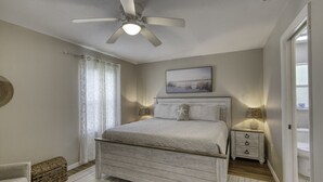 A bedroom with a bed, a ceiling fan, a nightstand, a lamp, a painting, and a door. - Last Mango in Palm Coast (Palm Coast)